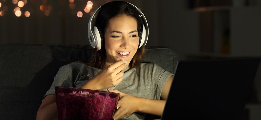 Frau mit Kopfhörern sitzt lächelnd mit Popcorn vor dem Bildschirm