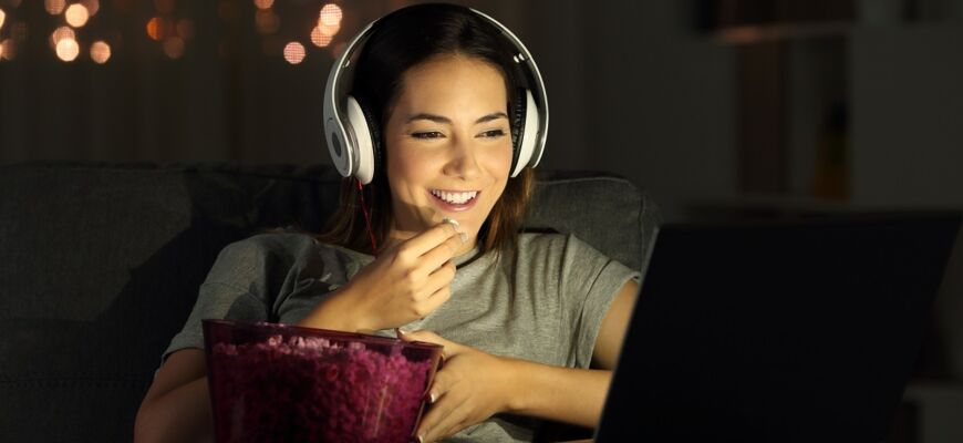 Frau mit Kopfhörern sitzt lächelnd mit Popcorn vor dem Bildschirm